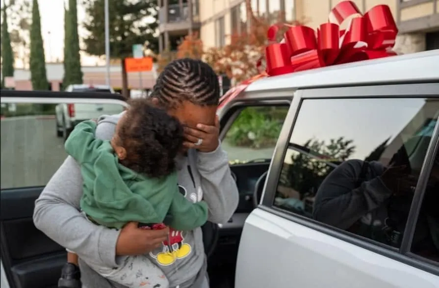 A woman holding a toddler and covering her eyes beside a white vehicle topped with a large red bow